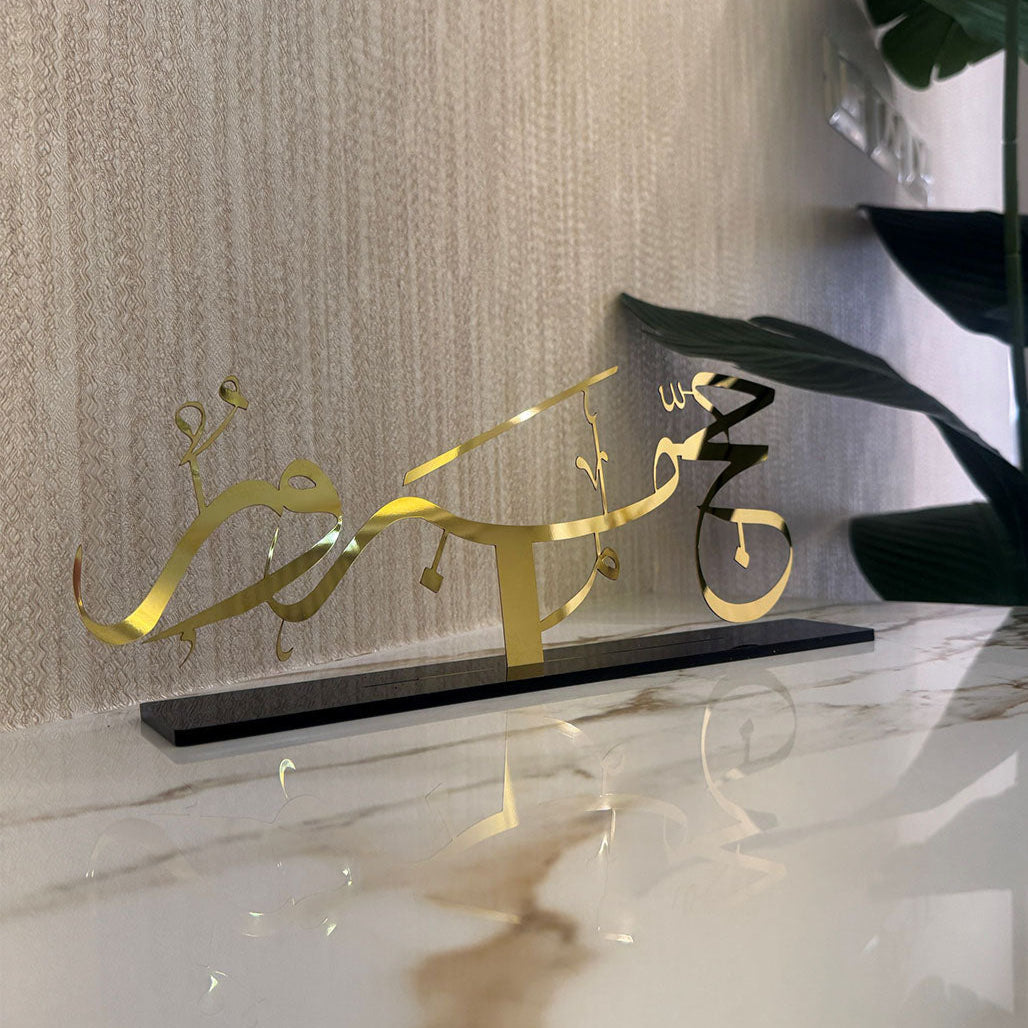 Decorative gold Arabic calligraphy sculpture on a marble surface with a textured wall background.