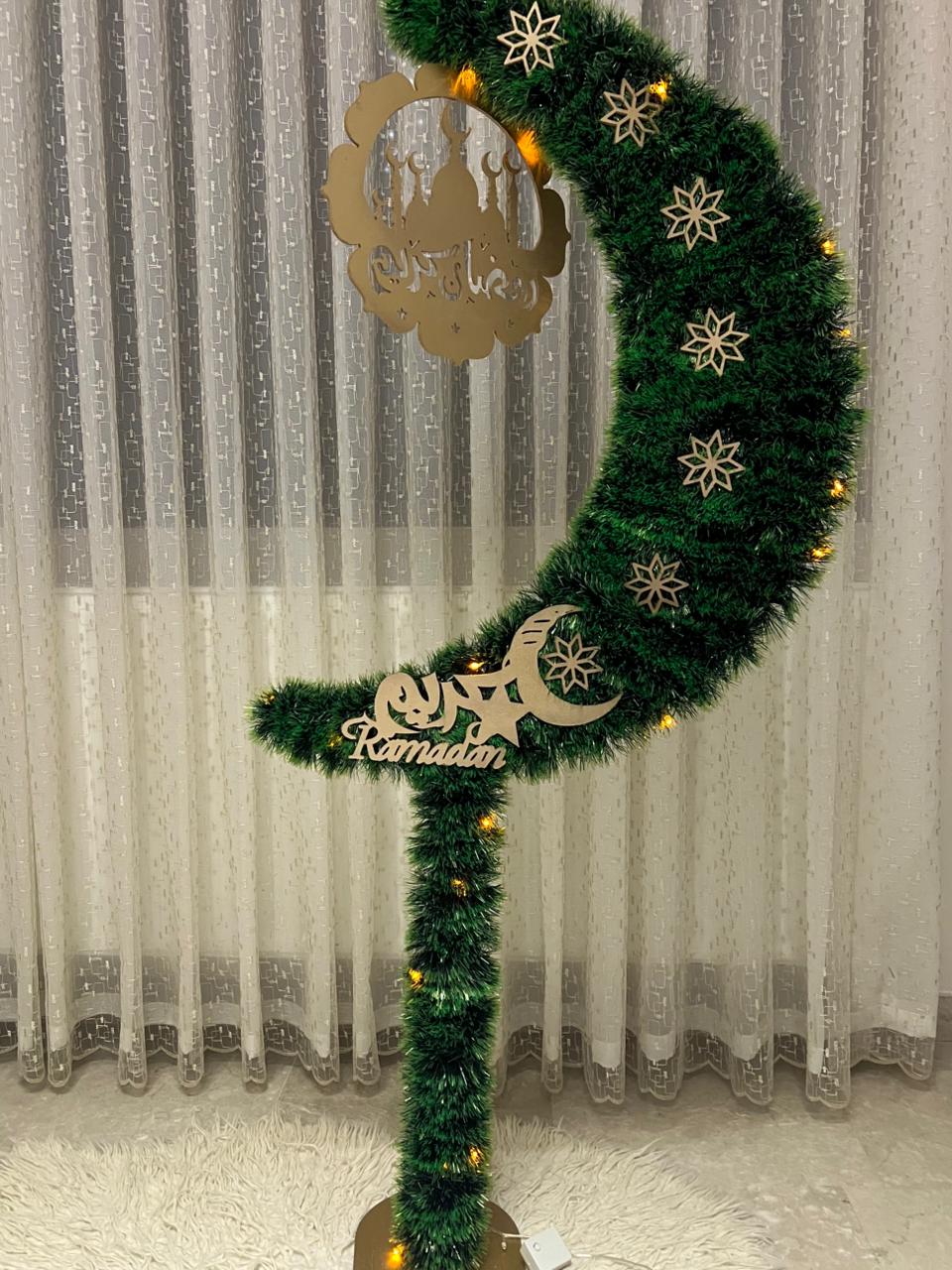 Decorative green crescent moon wreath with lights and 'Ramadan' sign against a curtain background