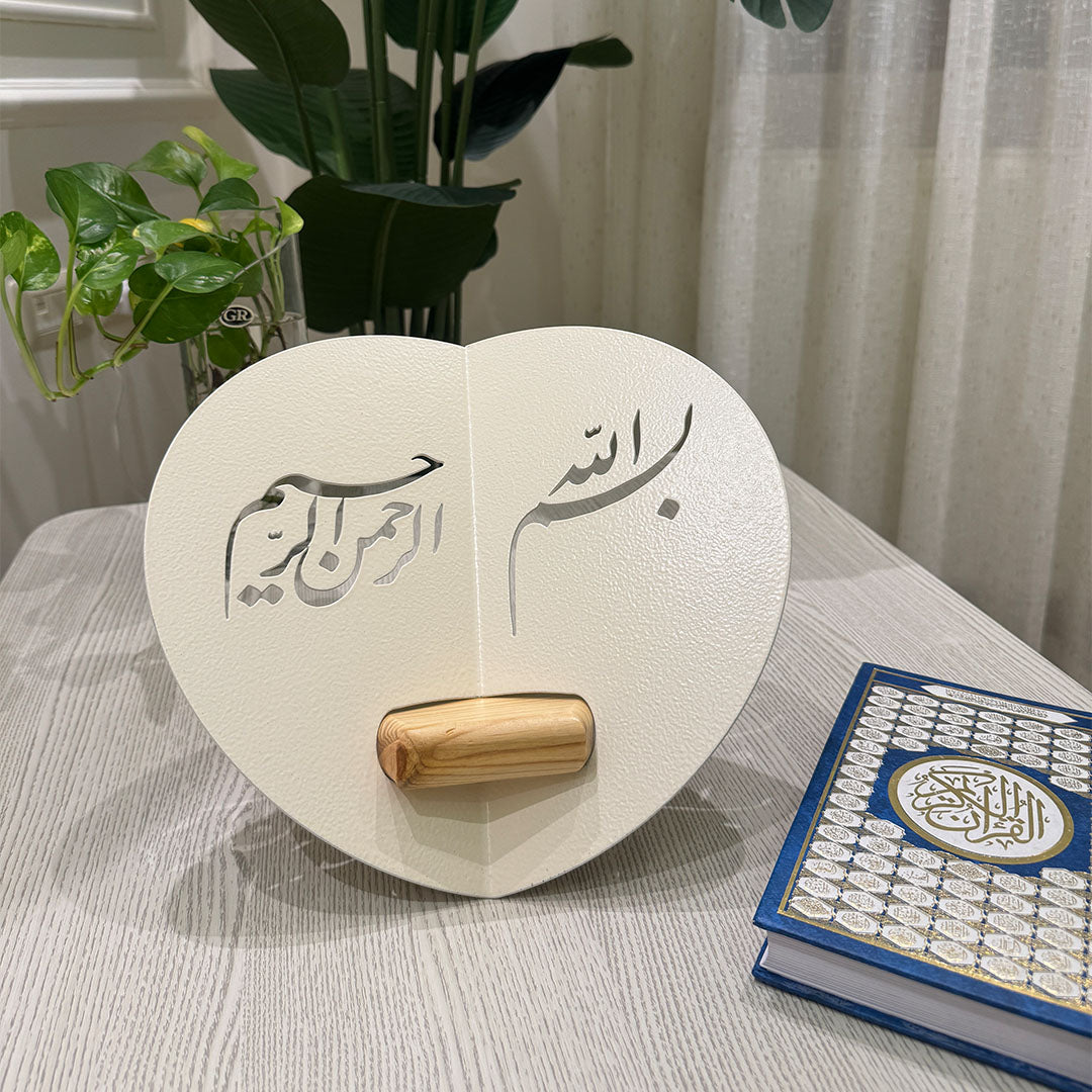 Heart-shaped wooden stand with Arabic calligraphy next to a Quran on a table.