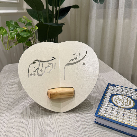 Heart-shaped wooden stand with Arabic calligraphy next to a Quran on a table.